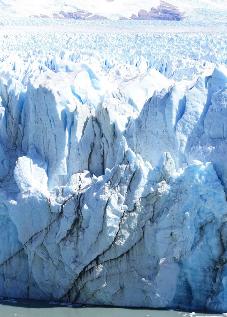 Dramatic close-up photograph of a massive blue glacial ice wall showing texture and melt, with mountains in the distance