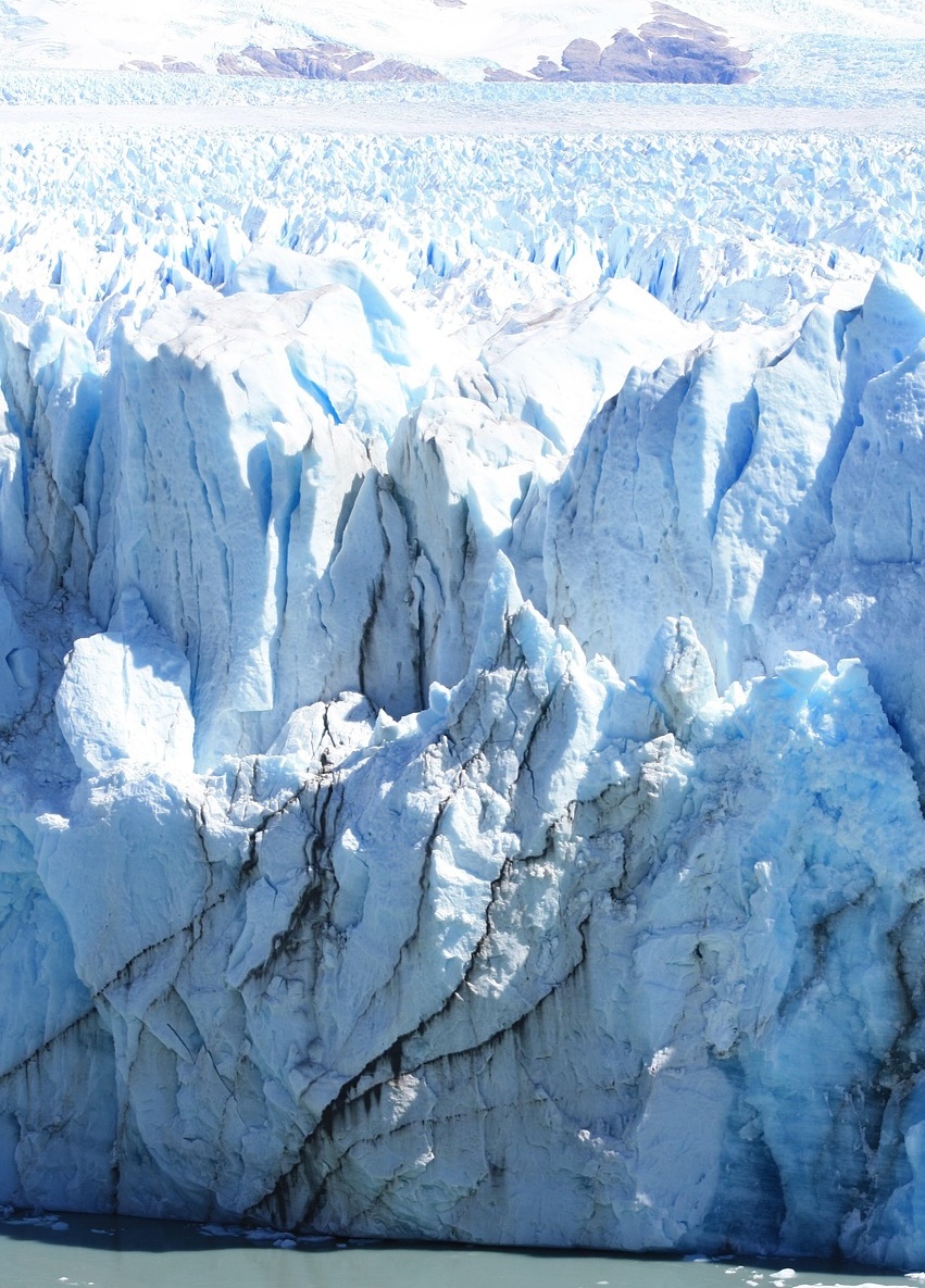 Dramatic close-up photograph of a massive blue glacial ice wall showing texture and melt, with mountains in the distance